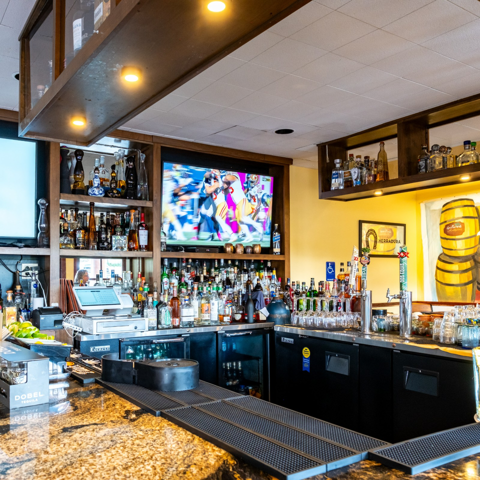 Well-stocked restaurant bar with liquor bottles, beer taps, sports television, granite counter, and tequila barrel artwork.