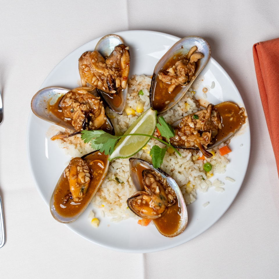 Six saucy mussels arranged over vegetable fried rice, garnished with lime wedges and fresh cilantro on a white plate.
