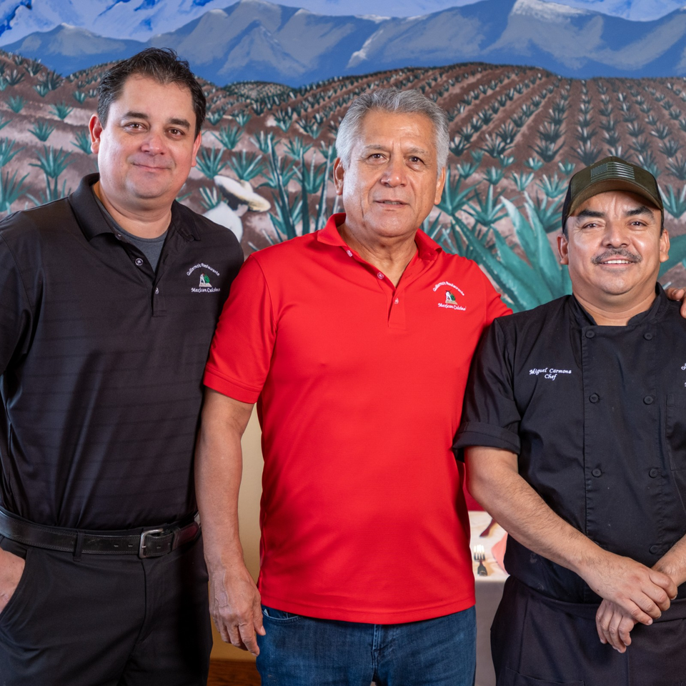 Three Mexican restaurant team members pose together before a mural depicting agave fields and mountains.