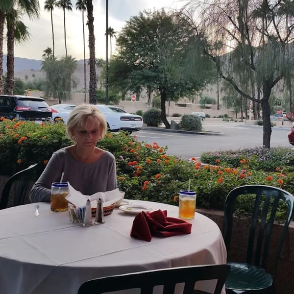 Woman seated at an outdoor table with drinks, overlooking a parking lot and palm trees.