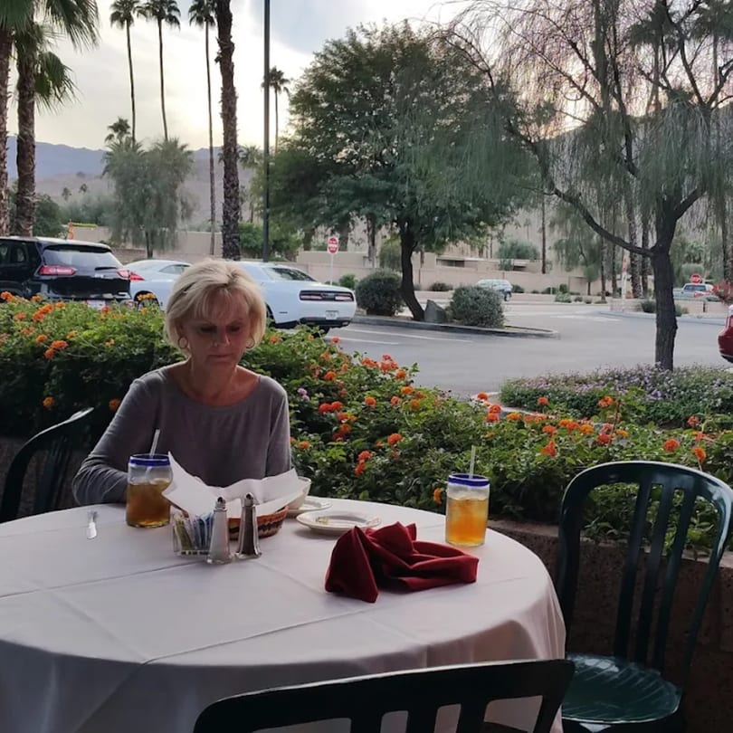Woman seated at an outdoor table with drinks, overlooking a parking lot and palm trees.