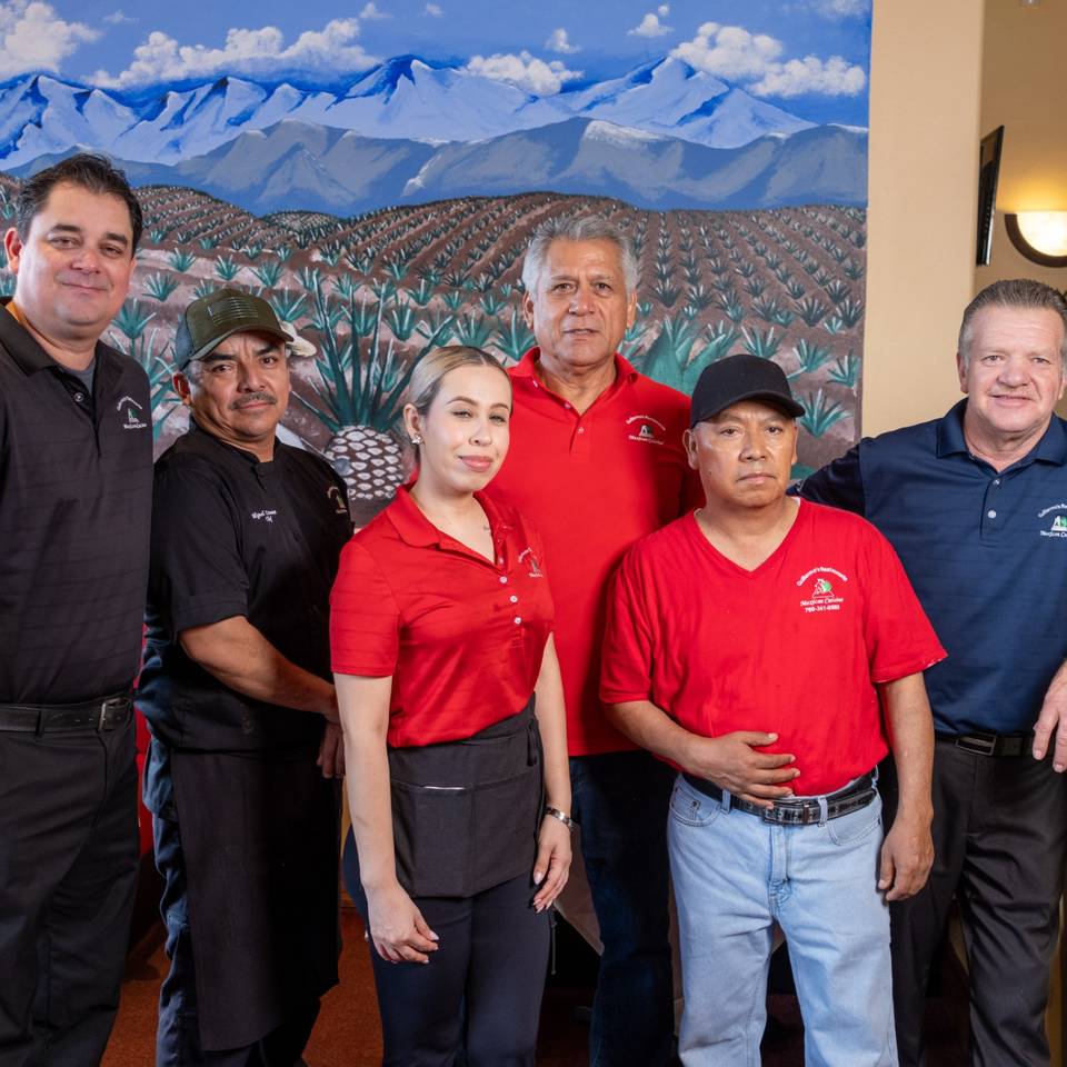 Six Mexican restaurant staff members pose together in front of a mural depicting agave fields and blue mountains.