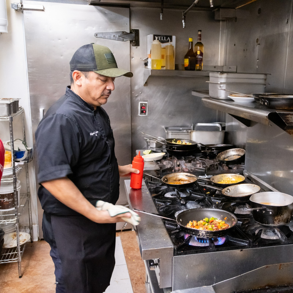 Restaurant chef prepares colorful vegetables in a pan over a gas stove inside a stainless-steel commercial kitchen.