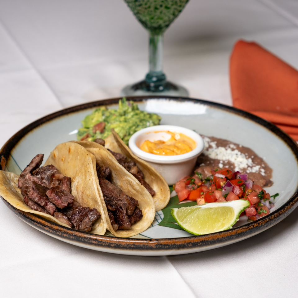 Plate of grilled steak tacos with guacamole, refried beans, queso, pico de gallo, lime, and orange sauce.