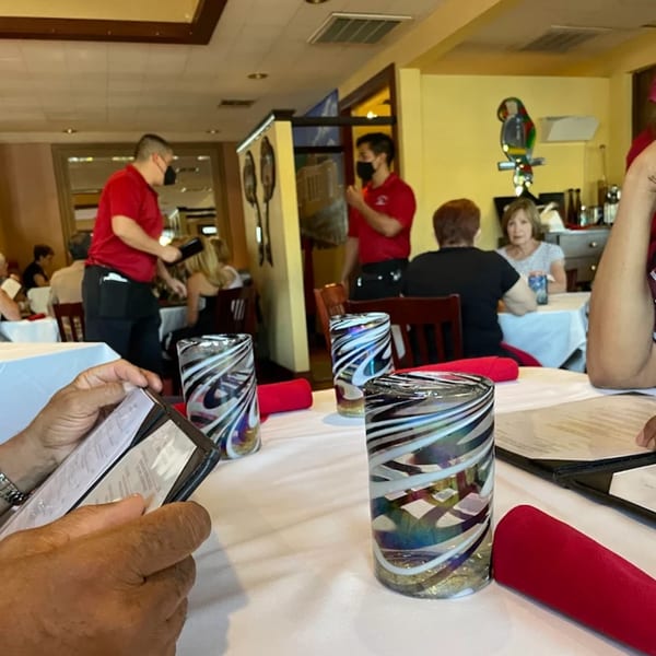 Diners seated at tables with white tablecloths and colorful glasses, with masked servers in red shirts.