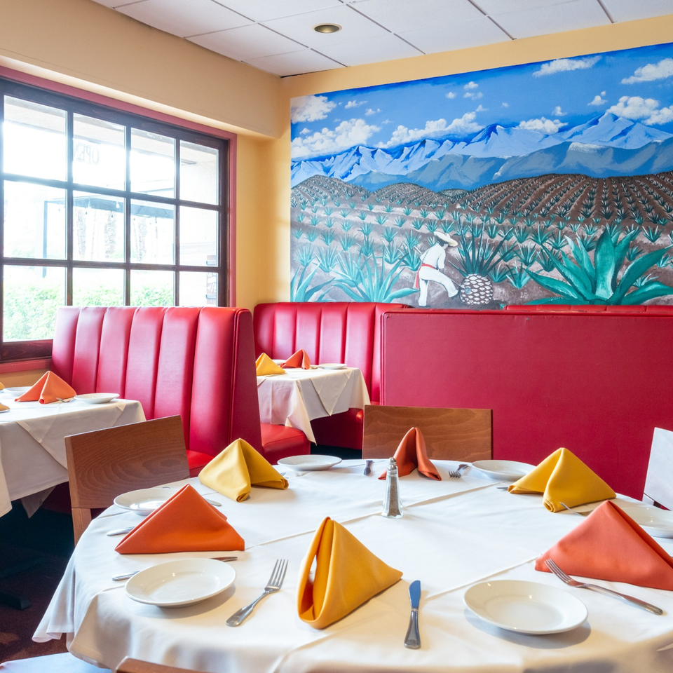 Restaurant dining room with red booths, white tablecloths, colorful napkins, and a mural of agave fields and mountains.