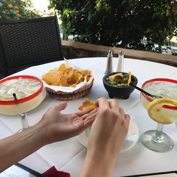 A person dips a tortilla chip into guacamole at an outdoor table with margaritas.