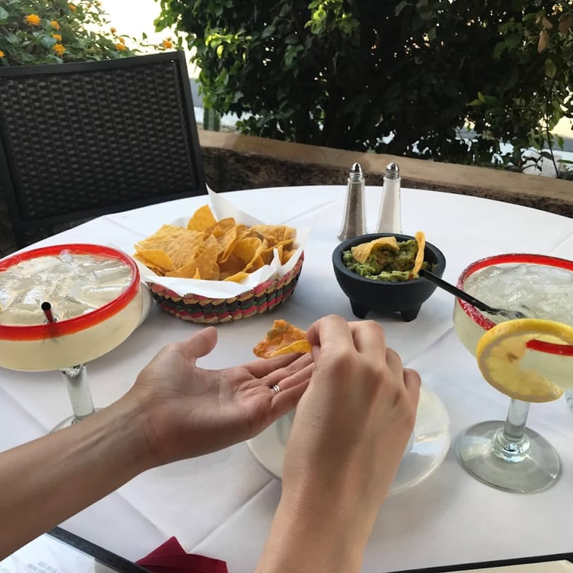 A person dips a tortilla chip into guacamole at an outdoor table with margaritas.