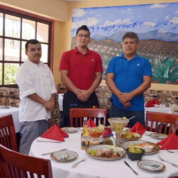 Three men, including a chef, stand proudly behind a table laden with Mexican dishes and drinks.