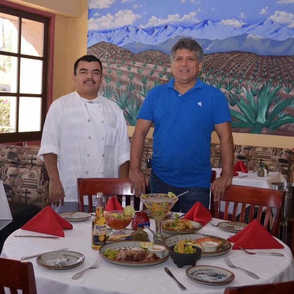 Two men, one in a chef's coat, stand behind a table laden with Mexican food and drinks.
