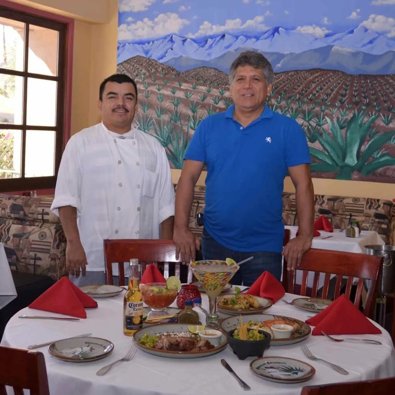 Two men, one in a chef's coat, stand behind a table laden with Mexican food and drinks.
