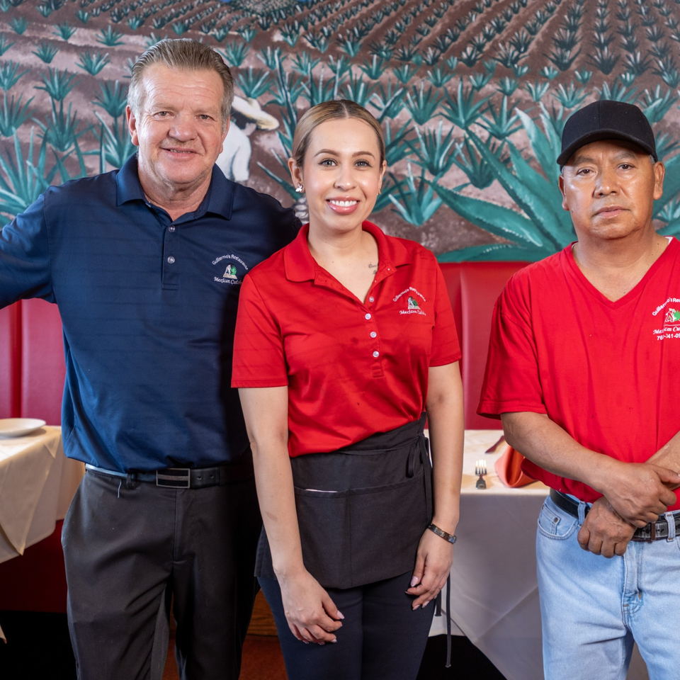 Three restaurant staff members pose together in front of agave-themed artwork and dining tables.