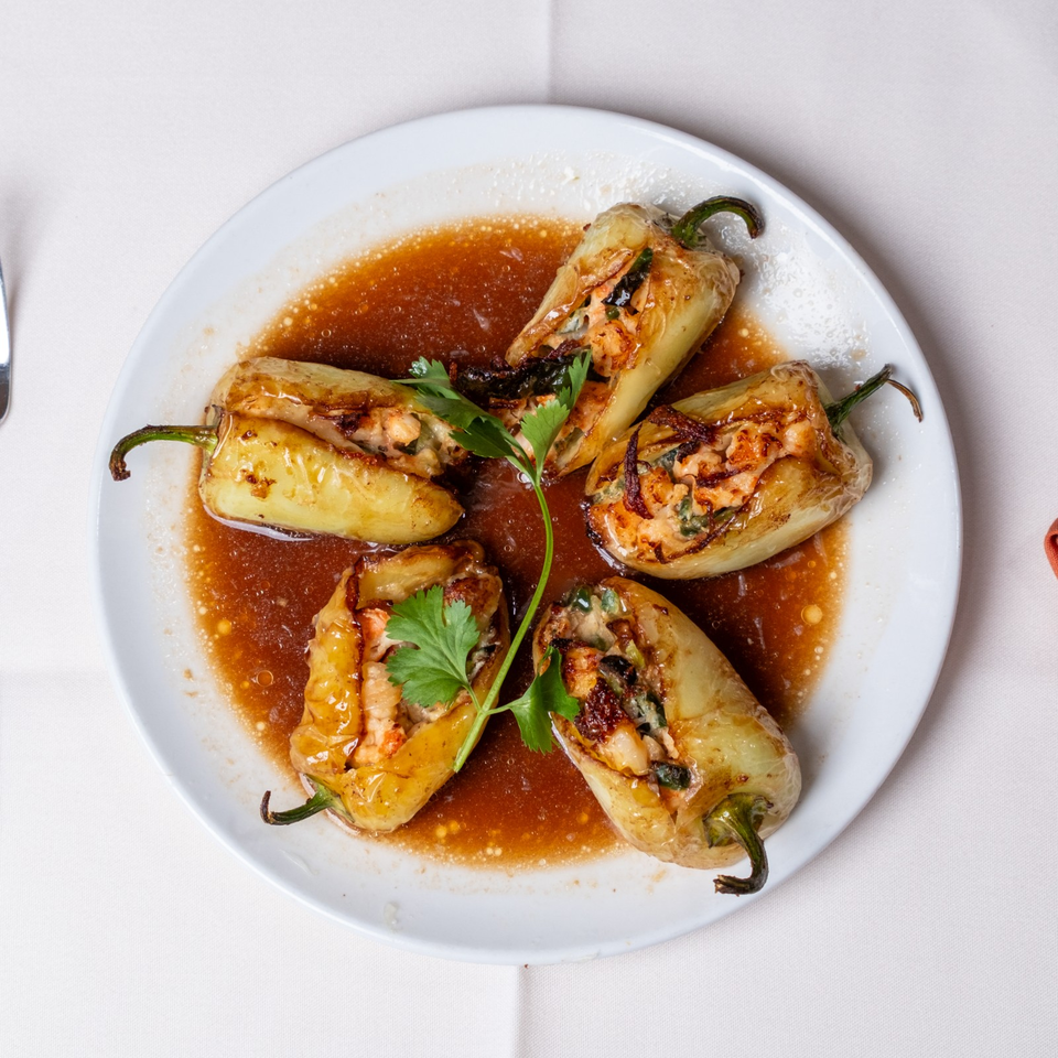 Four roasted stuffed peppers arranged on a white plate with savory brown sauce and fresh parsley garnish.