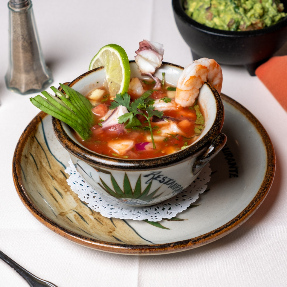Seafood ceviche with shrimp, octopus, lime, cilantro, and vegetables served in a ceramic bowl with guacamole in the background.